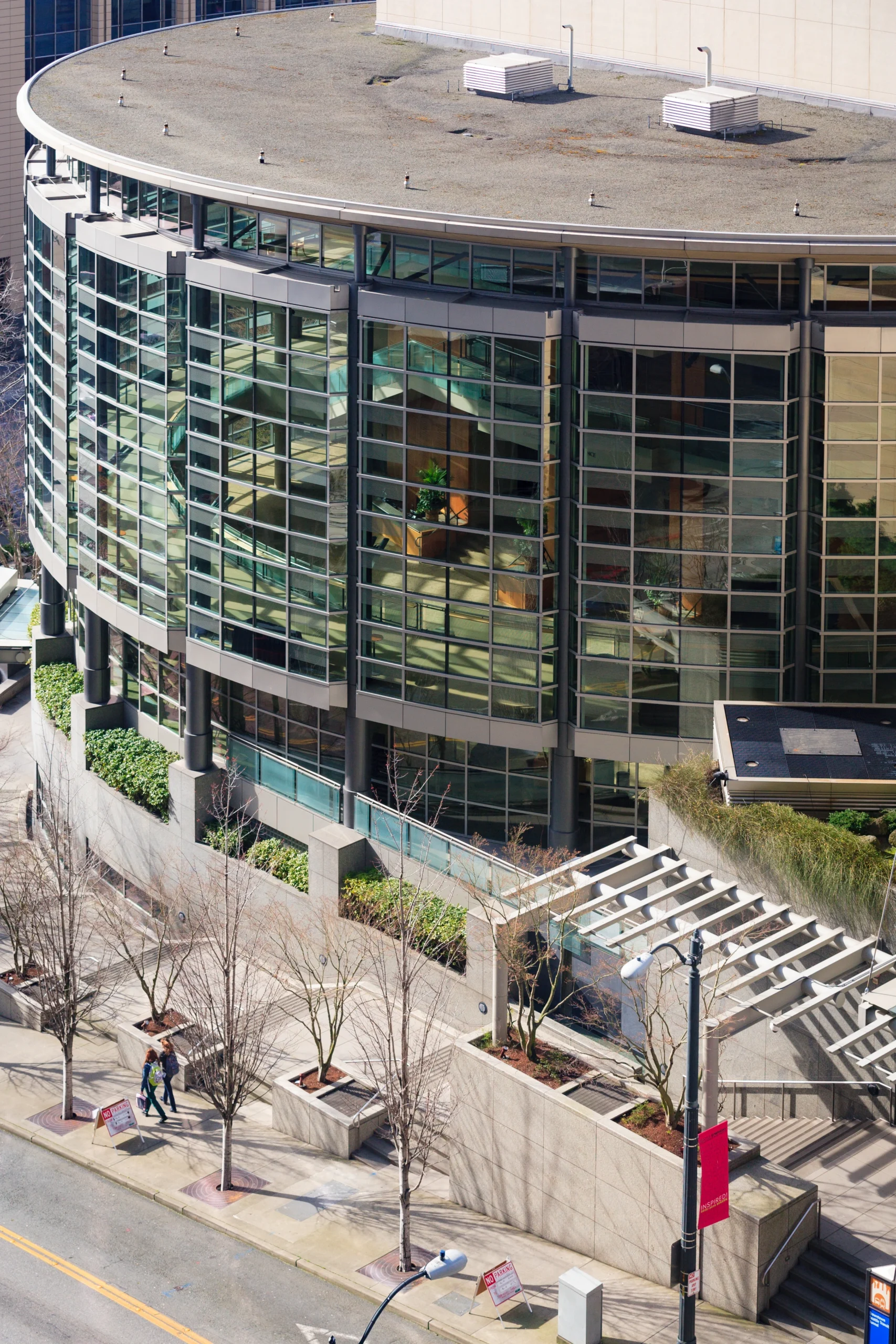 Aerial view of a curved glass office building with multiple windows, rooftop HVAC units, and a sidewalk with leafless trees below.