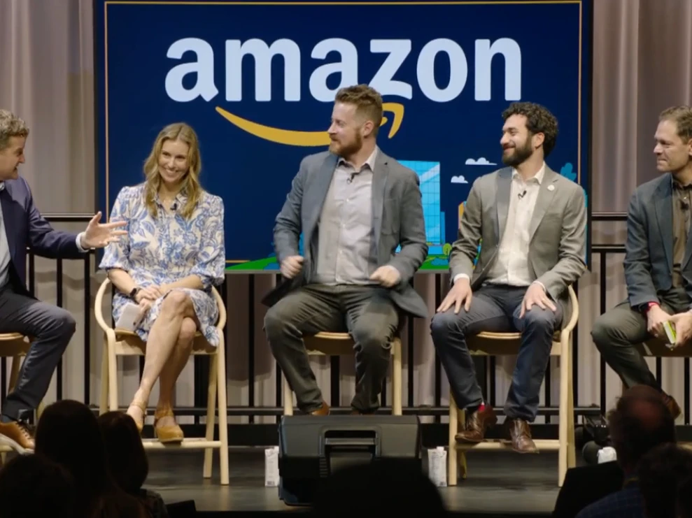 Panel discussion on a stage with five speakers seated on stools in front of a large Amazon sign.