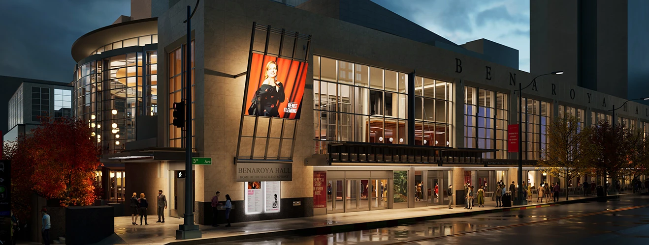 Night view of Benaroya Hall exterior with glass frontage, a large performer poster, and pedestrians along a wet street.