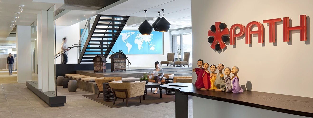 Contemporary office lobby with a black staircase, lounge seating, world map wall art, and a red PATH logo near colorful figurines on the counter.