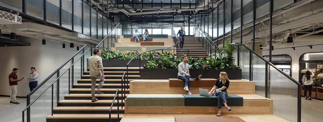 Open two-story atrium with tiered wooden seating, glass railings, and lush planters as people work on laptops and chat.