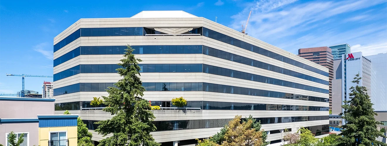 Modern office building with horizontal beige and dark blue stripes under a clear blue sky, surrounded by trees and neighboring structures
