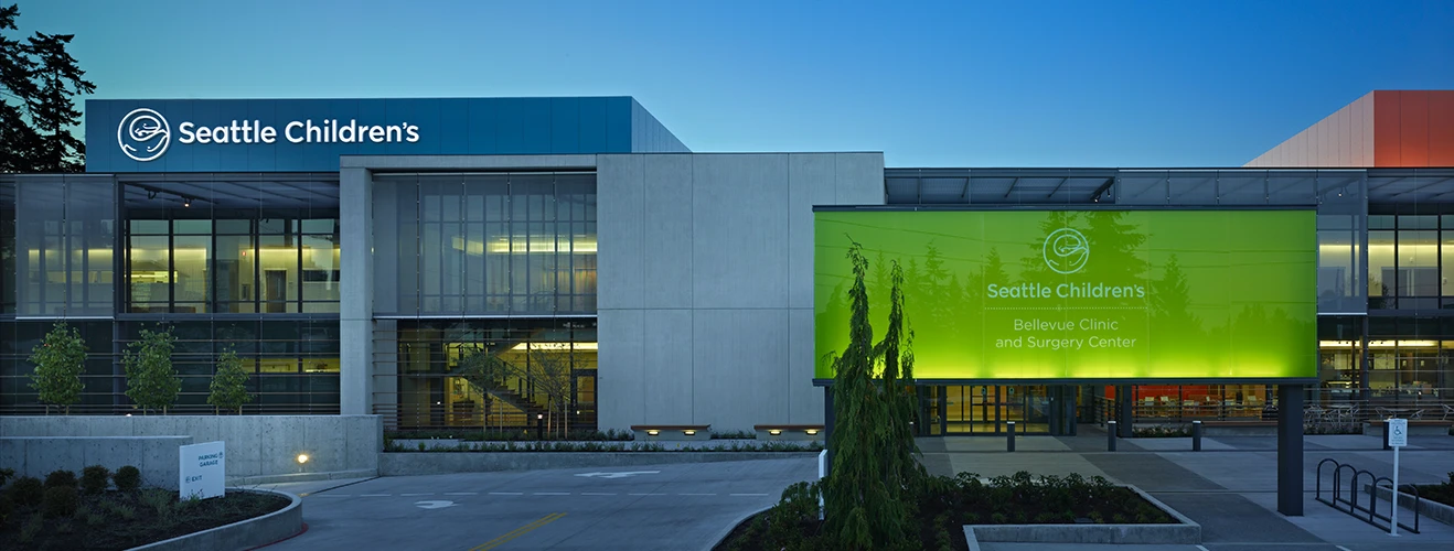 Exterior view of Seattle Children's campus with a blue sign and glass-fronted building, and a large green banner advertising Bellevue Clinic and Surgery Center.