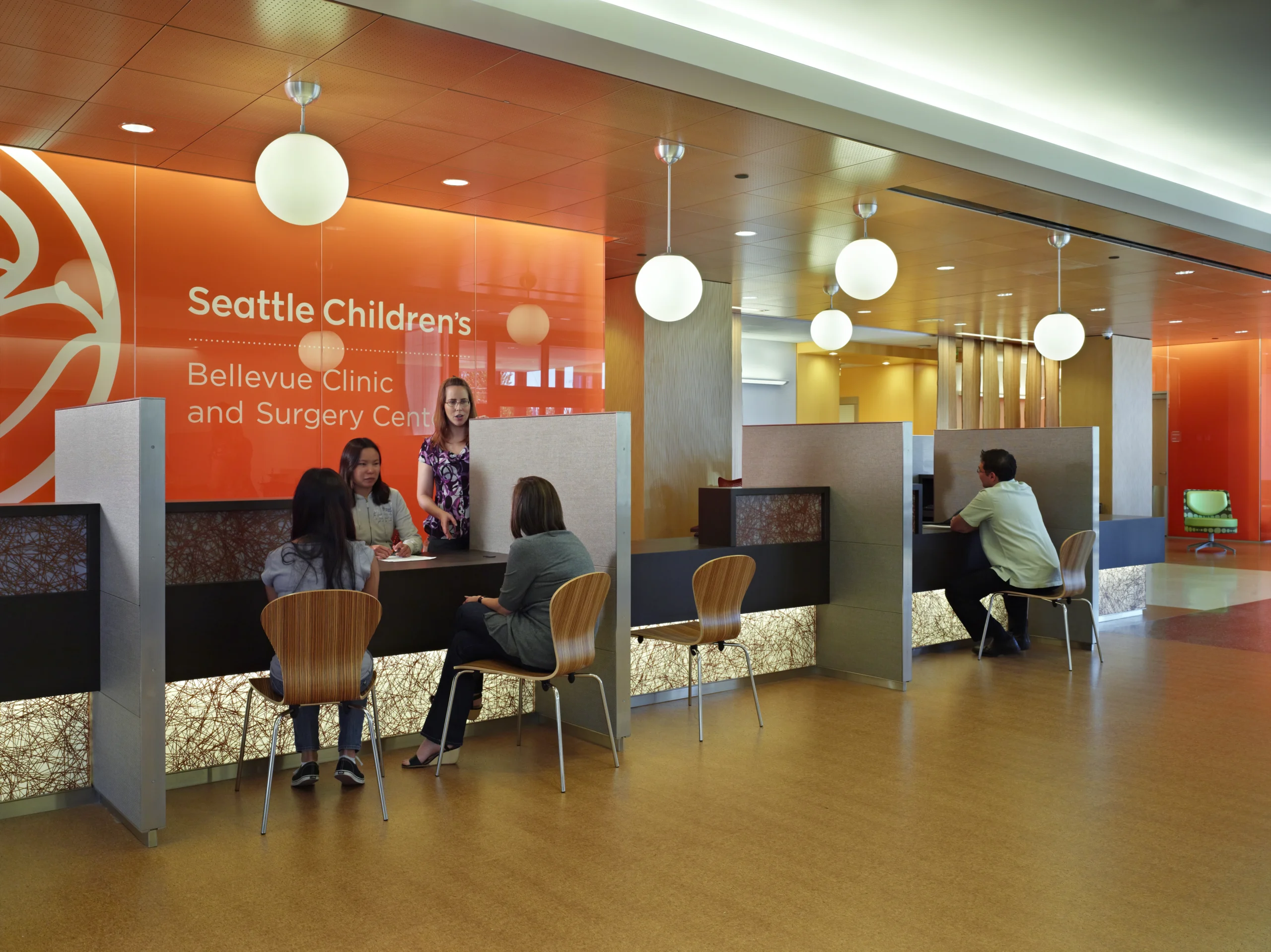 Reception area at Seattle Children's Bellevue Clinic and Surgery Center with an orange backdrop, staff at desks, and families seated nearby.