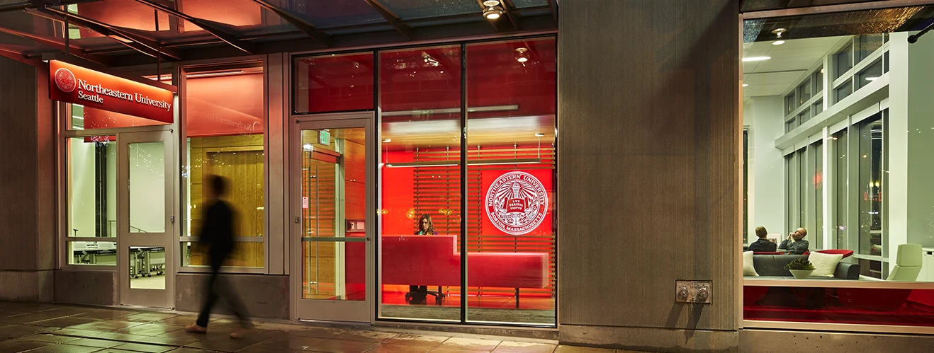 Exterior view of Northeastern University Seattle at night, with glass doors, a red-lit reception area, and the university seal visible inside. A blurred person walks along the sidewalk in front of the entrance.